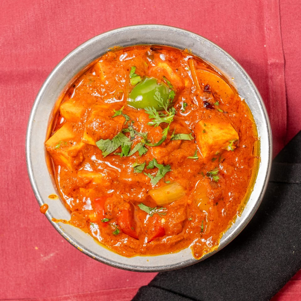Overhead view of a metal bowl filled with vibrant orange paneer curry, garnished with cilantro and bell peppers.