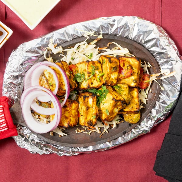 Sizzling paneer tikka with onions and cabbage on a hot plate, served on a red tablecloth.