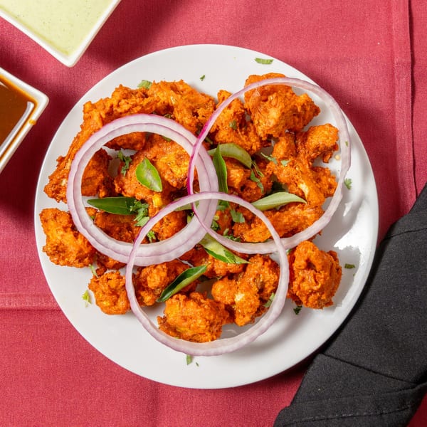 Crispy orange chicken pakora with red onion rings and curry leaves on a white plate.