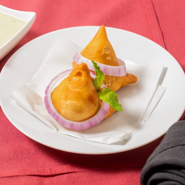 Two golden-brown samosas garnished with red onion rings and mint on a white plate.