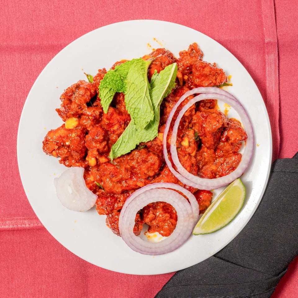 Spicy red chicken dish garnished with mint, red onion rings, and a lime wedge on a white plate.